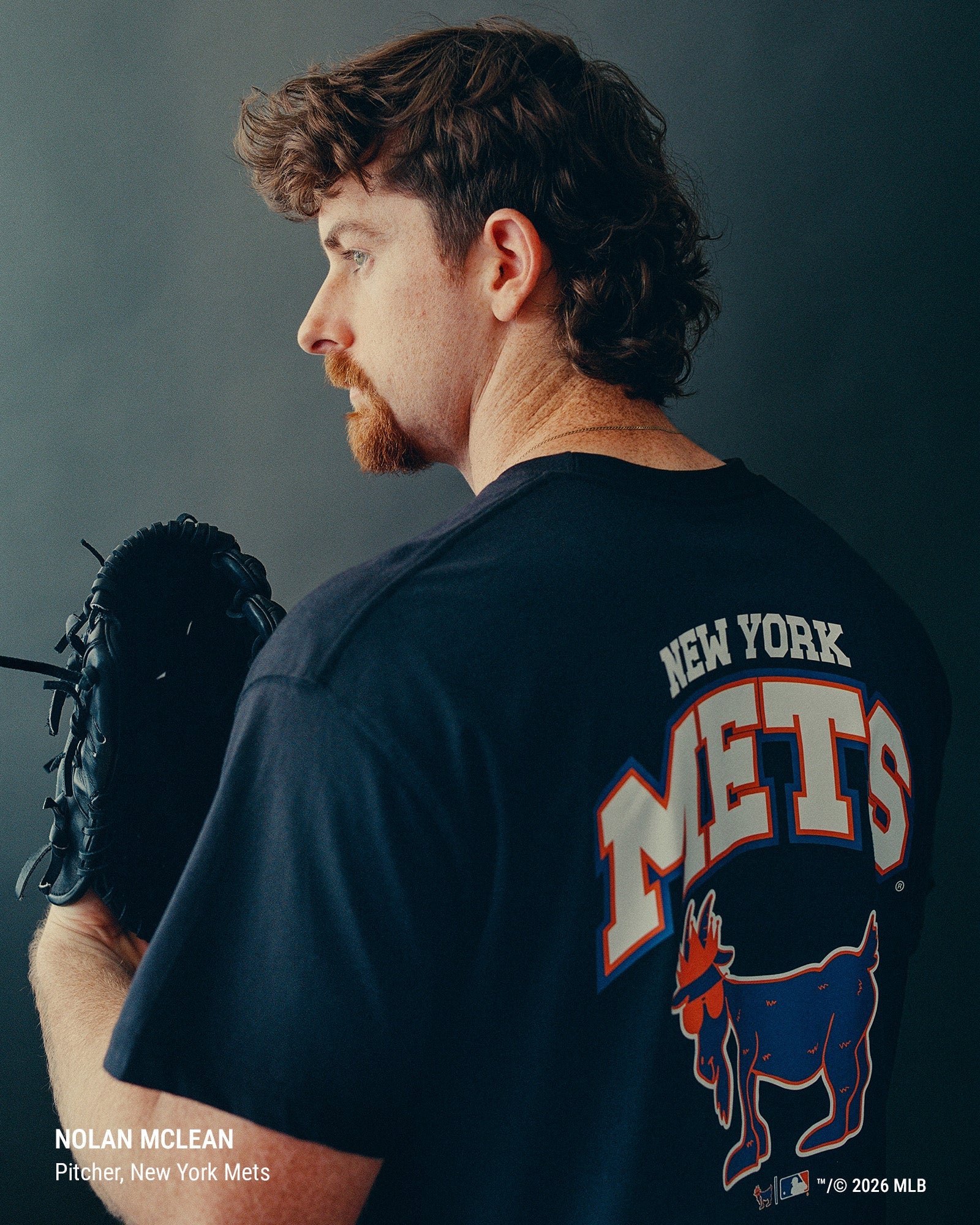 A man shown from the back wearing a dark navy New York Mets graphic T-shirt, holding a black baseball glove against a dark studio background. The shirt features a large white, red, and blue Mets logo on the back.#color_black