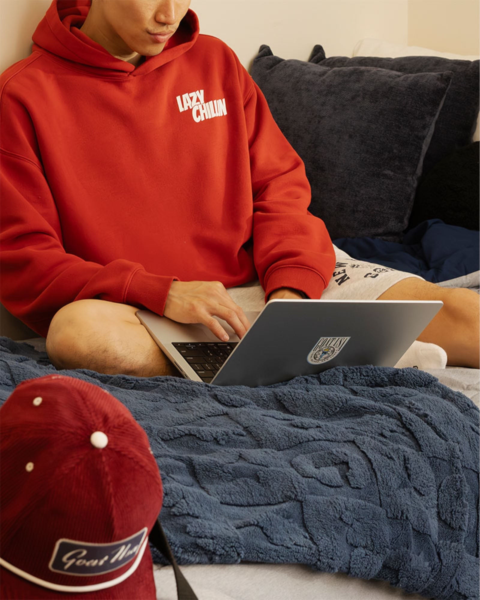 A person sitting on a bed wearing a bright red hoodie, using a laptop. A navy blue textured blanket covers the bed, and a red baseball cap is visible in the foreground.