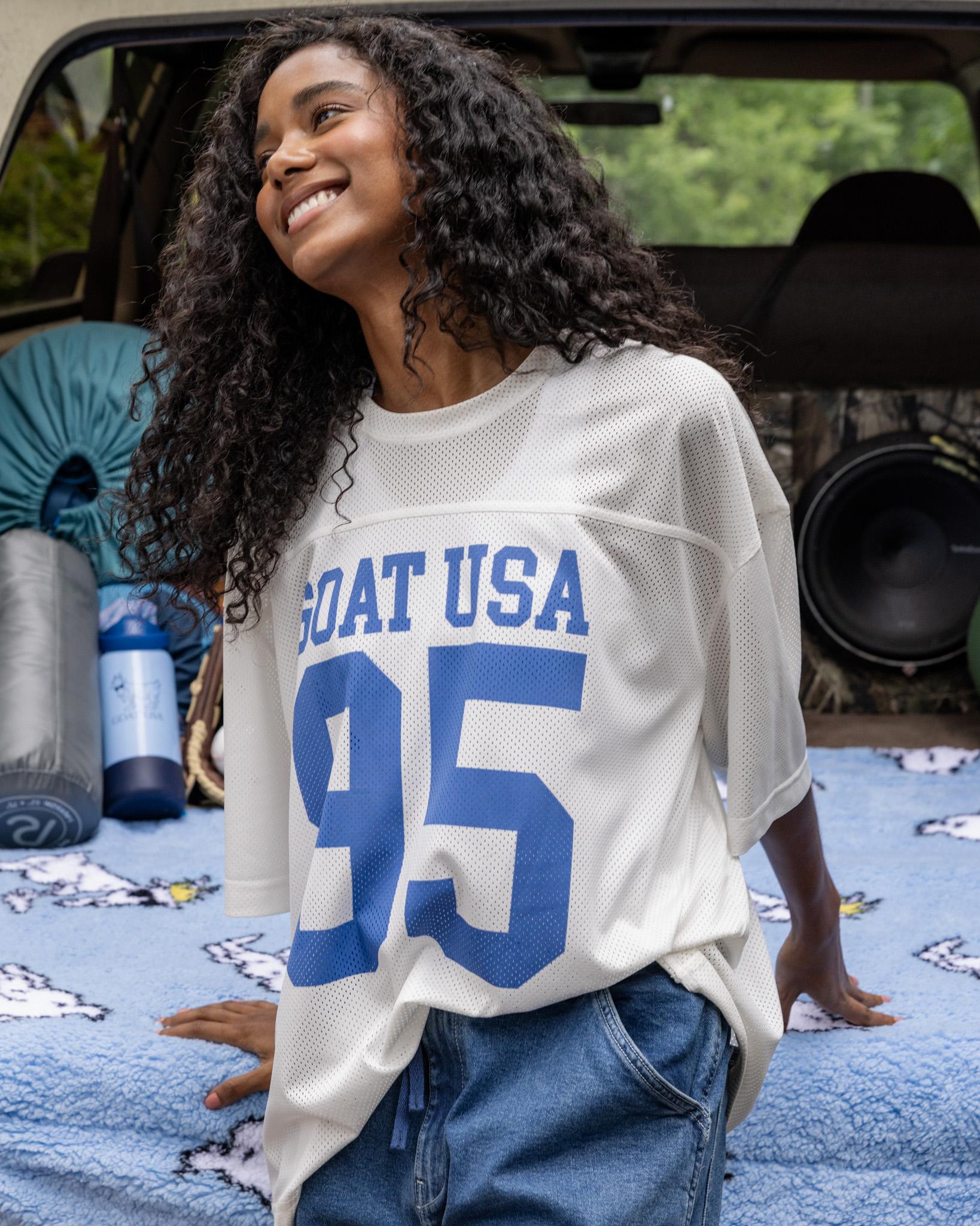 Woman wearing a white sports jersey with blue text and numbers, standing in front of a vehicle.