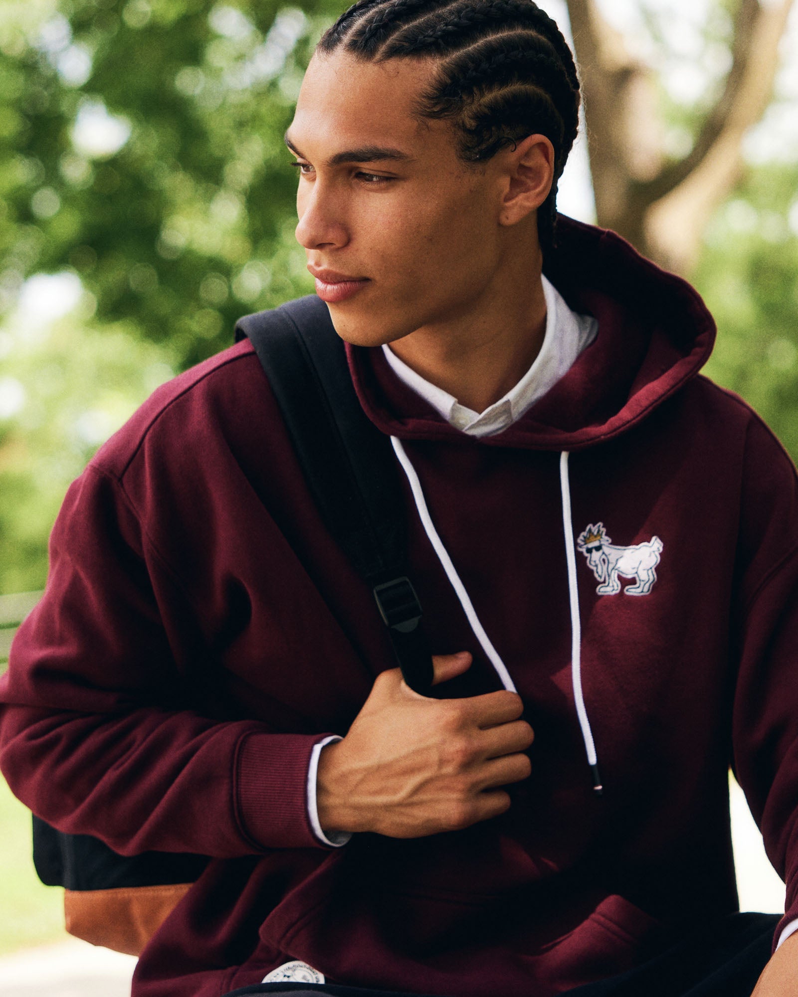 Man wearing a maroon hoodie with a logo, standing outdoors with greenery in the background #color_windsor-wine