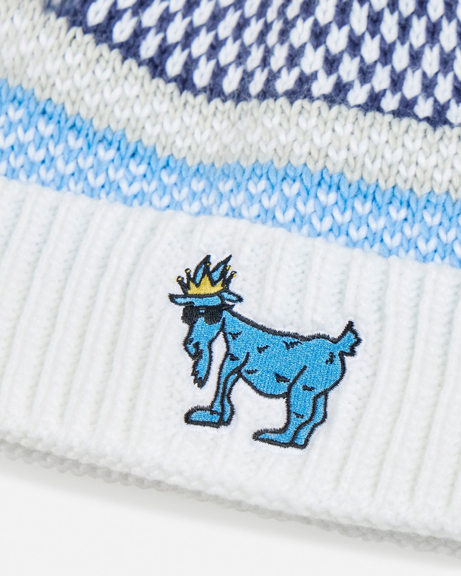 Close-up of a white knit beanie with blue and white pattern and embroidered blue goat logo.