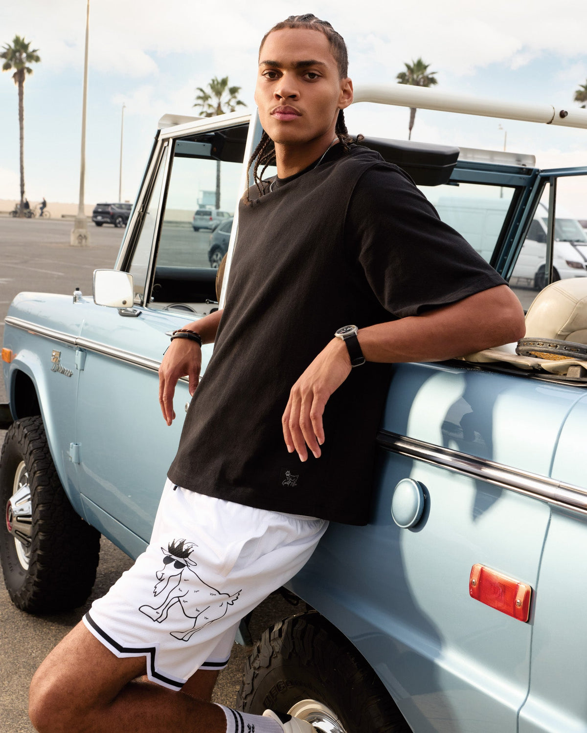 Person leaning against a vintage light blue truck with palm trees in the background