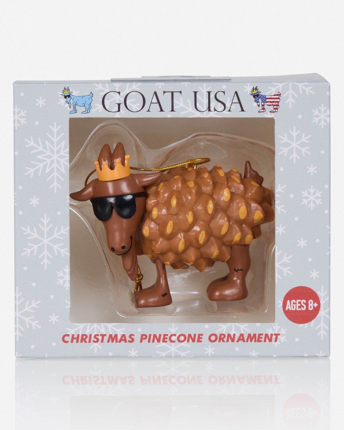 A Christmas pinecone ornament shaped like a goat wearing sunglasses and a gold crown, displayed inside its retail packaging labeled 'GOAT USA'.