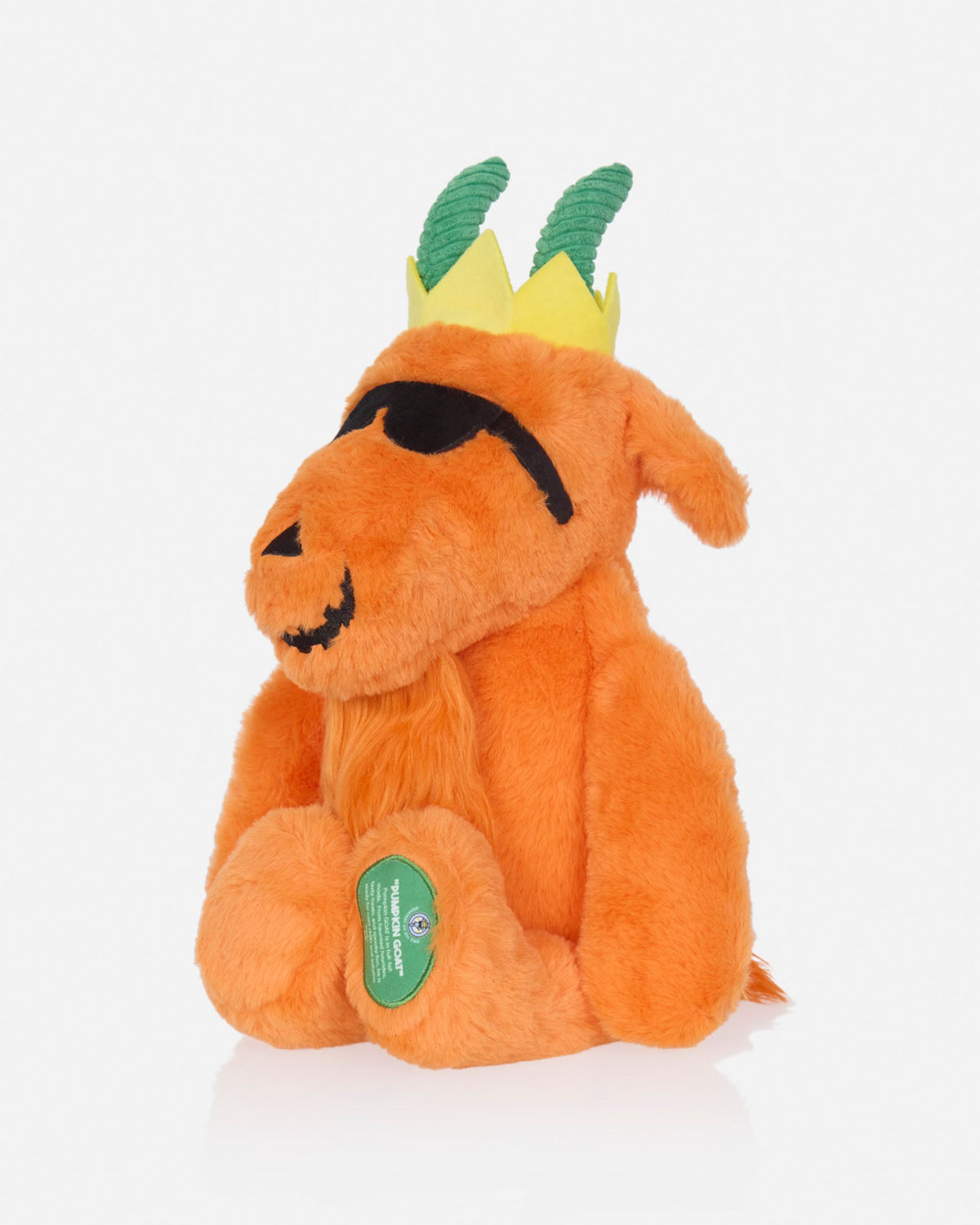 An orange plush goat toy with a yellow and green crown, black embroidered eyes and mouth, and a green circular tag on its front paw, photographed on a clean white background.