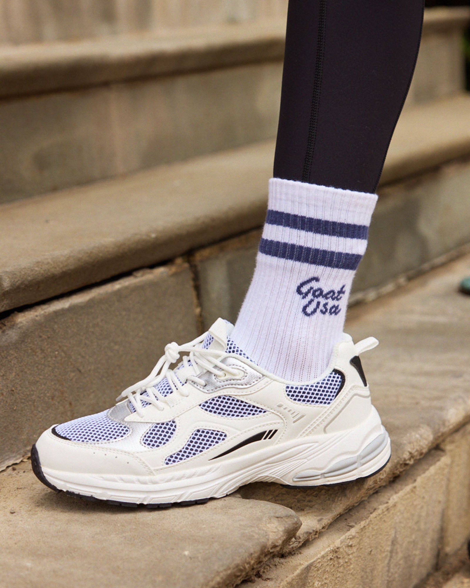 White sneakers with blue accents on a step, wearing white socks with blue stripes and 'Goat USA' branding.#color_navy