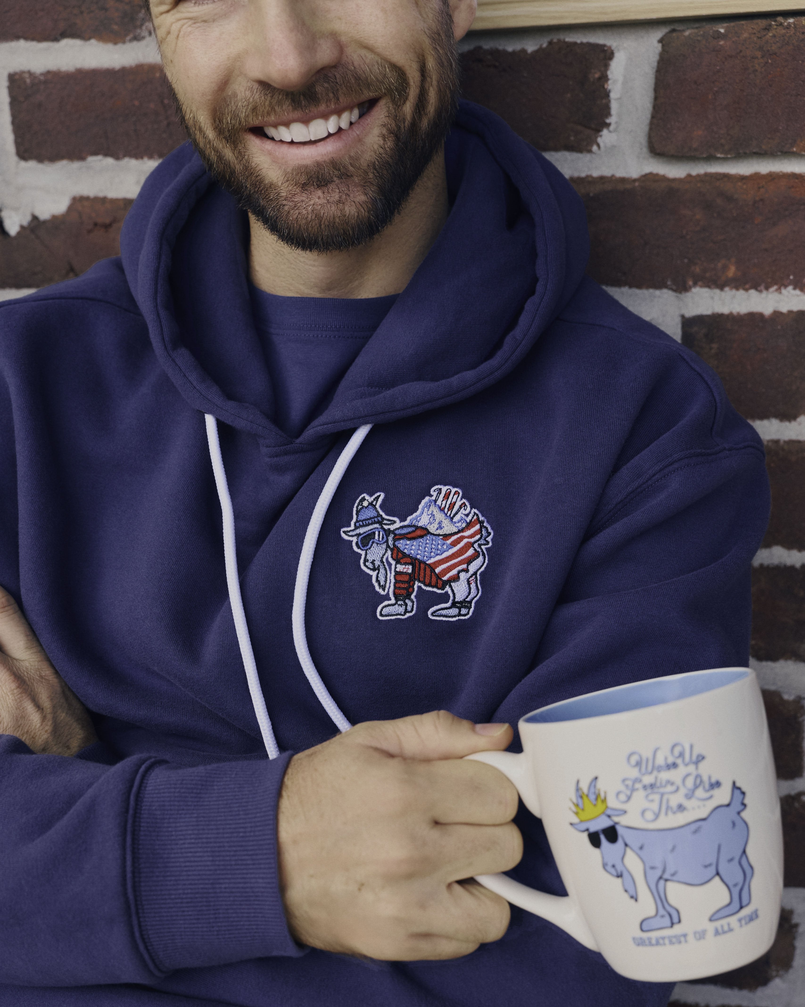 Man wearing a blue hoodie with a logo, holding a mug with a dog illustration, against a brick wall.