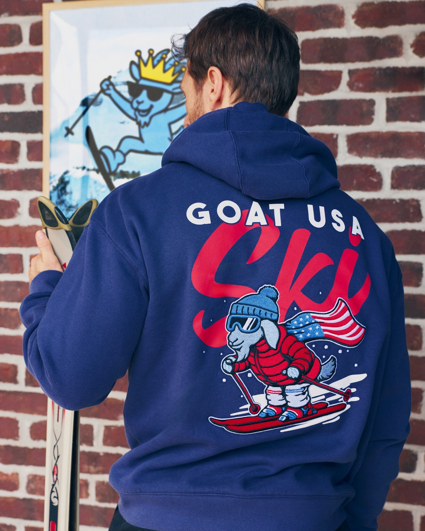 Person wearing a blue hoodie with a graphic design on the back, standing against a brick wall.