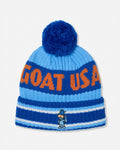 A blue and light blue knit beanie hat with a large blue pom-pom on top. The front has bold orange lettering that reads 'GOAT USA' and a small cartoon mascot embroidered near the bottom of the cuff.
