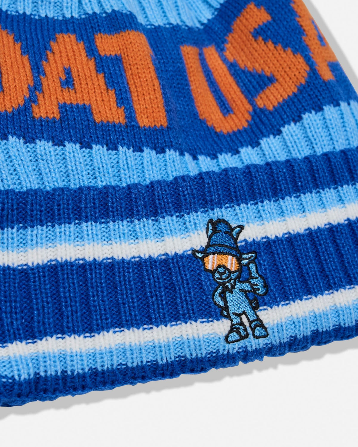 Close-up of a blue and light blue striped knit beanie with orange lettering and a small embroidered cartoon character on the brim.