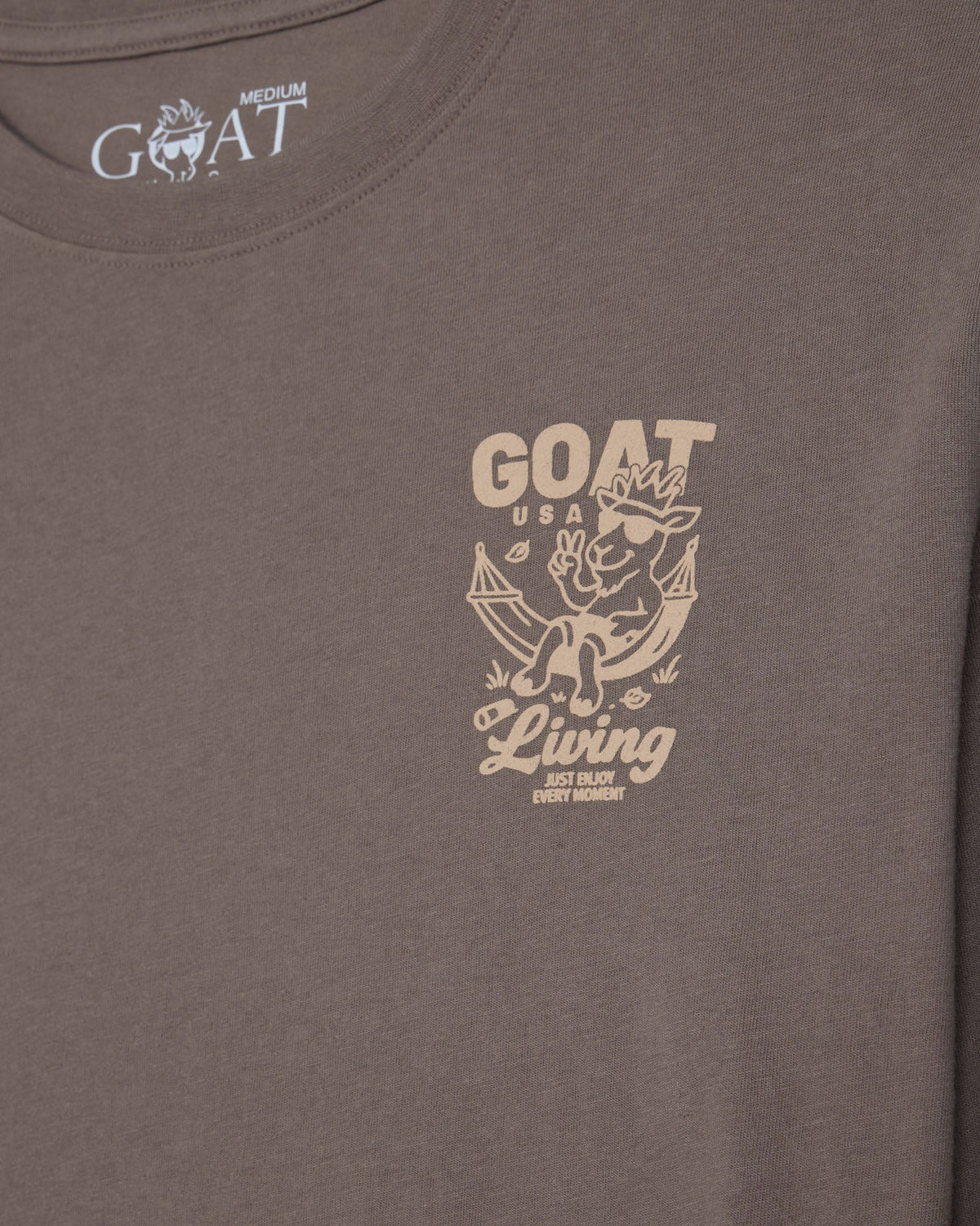 A close-up of a taupe brown t-shirt featuring a beige graphic print on the left chest with a cartoon goat and the text 'GOAT USA Living'. The inside collar shows the brand name 'GOAT' printed in white.