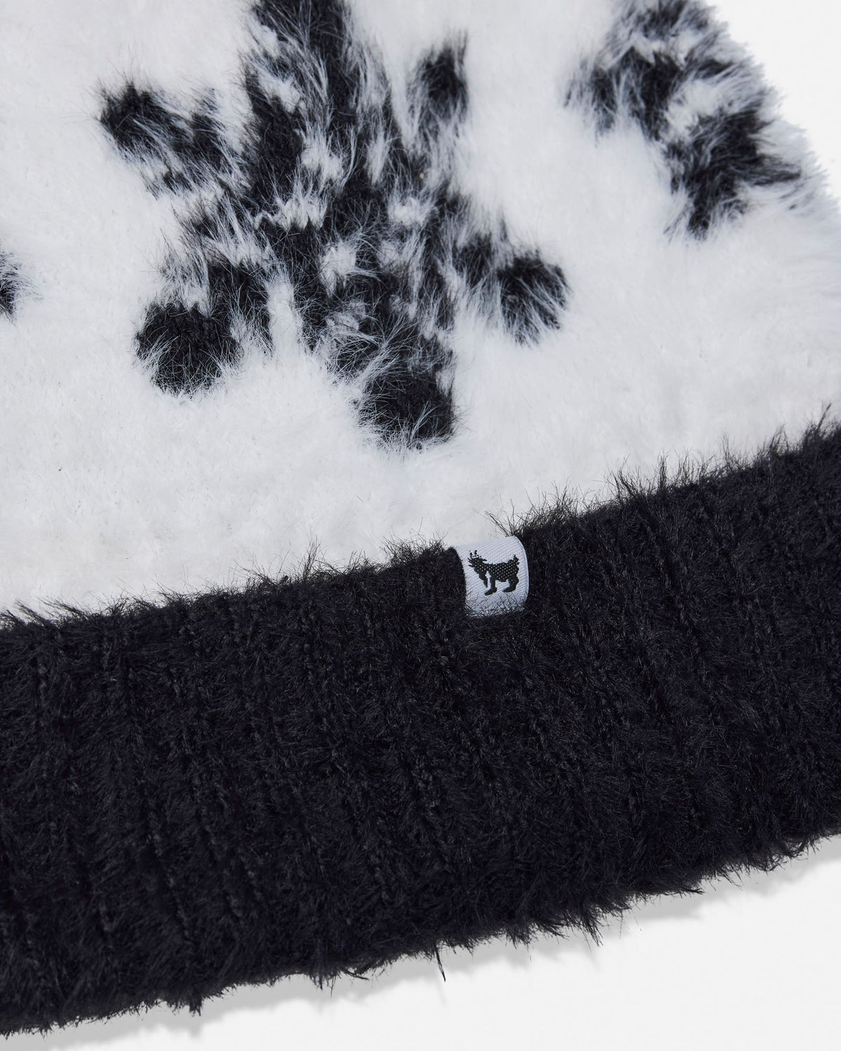 Close-up of a black and white fuzzy knit beanie with a ribbed black cuff and a small white tag featuring a black animal logo.