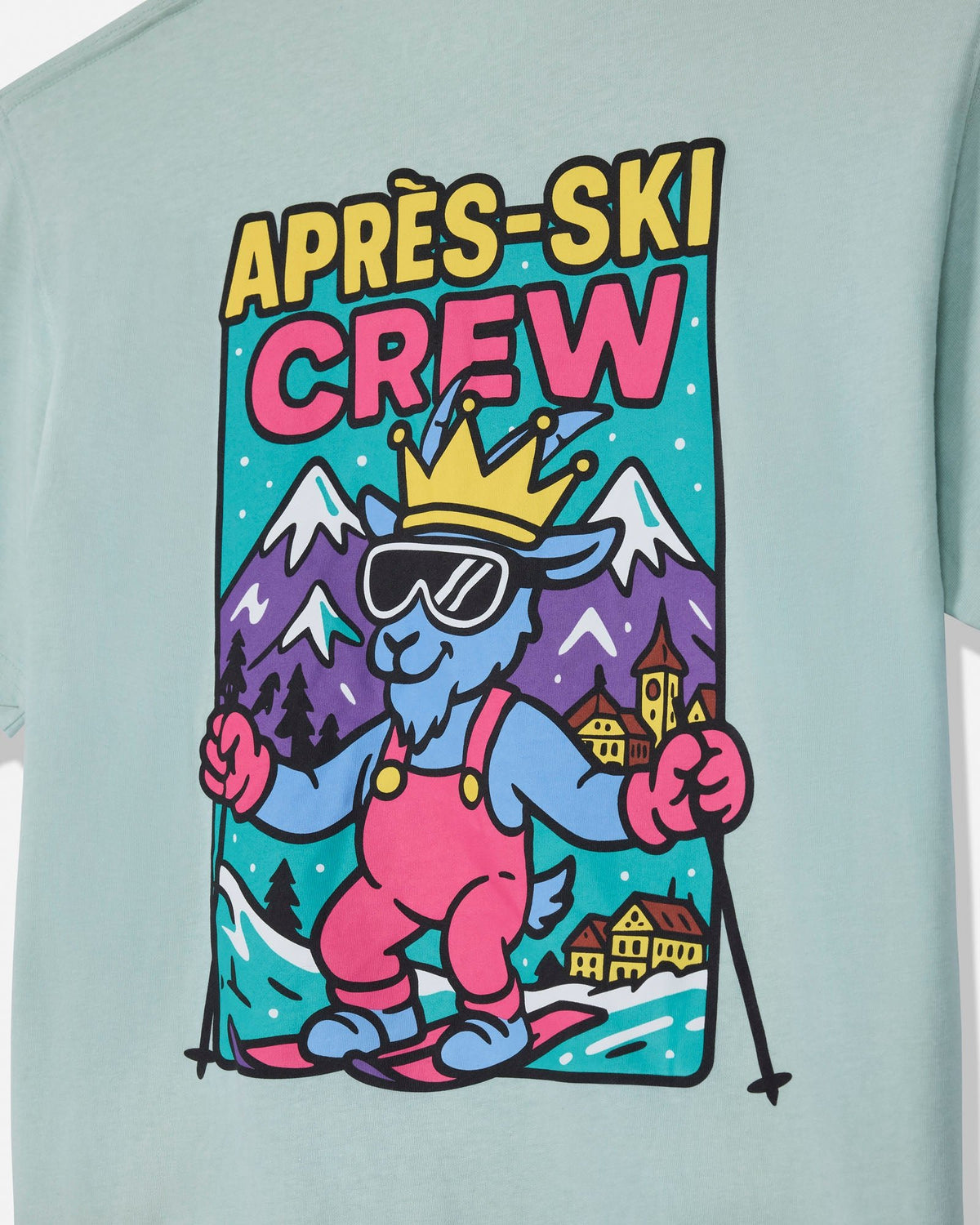 A close-up of a light mint green t-shirt featuring a colorful graphic print on the front. The graphic displays a cartoon blue goat wearing a gold crown, pink overalls, and blue ski boots, with the text 'APRÈS-SKI CREW' above it against a mountain background.
