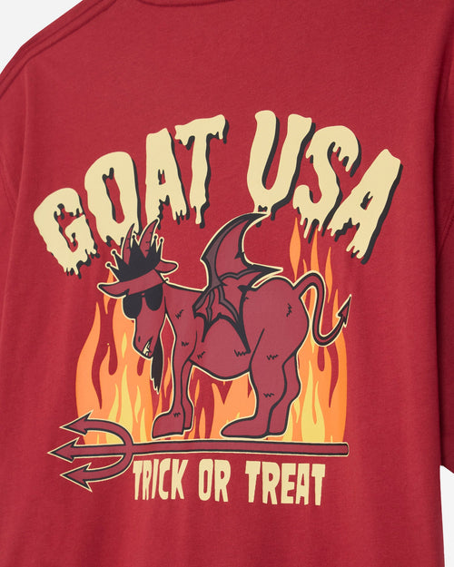A close-up of a red graphic t-shirt featuring a cartoon dragon goat wearing sunglasses and a hat, standing in front of flames. The text 'GOAT USA' is written above in a dripping cream font, and 'TRICK OR TREAT' is written below.