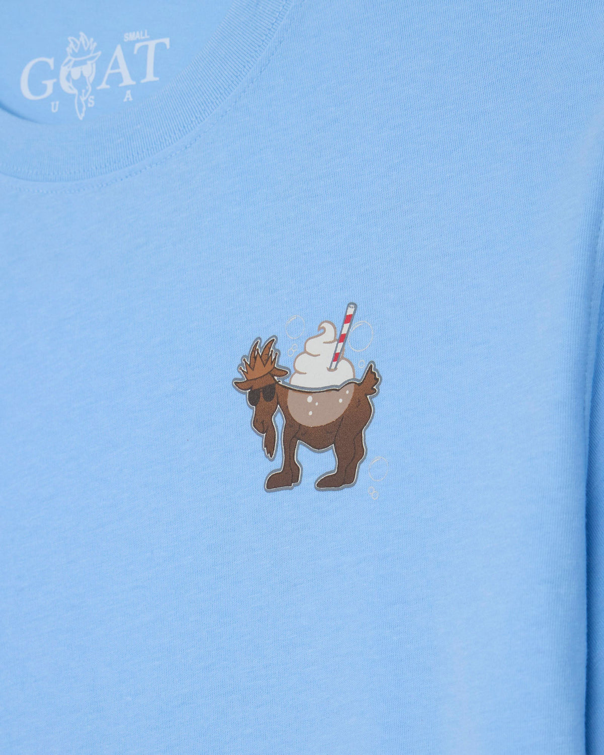 A close-up of a light blue t-shirt featuring a small graphic of a brown goat wearing sunglasses and holding a whipped cream sundae with a red-and-white striped straw. The brand name 'GOAT USA' is printed in white on the upper left chest.