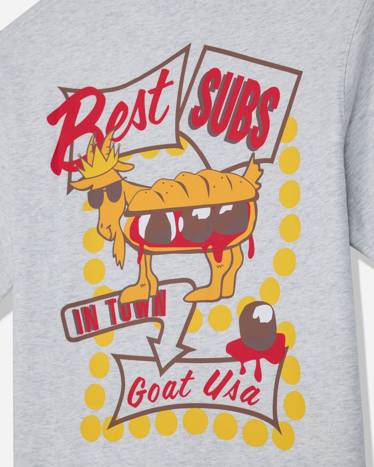 Close-up of a heather gray graphic T-shirt featuring a cartoon goat character with sunglasses and speech bubbles that read 'Best SUBS IN TOWN Goat USA' with yellow polka dots.