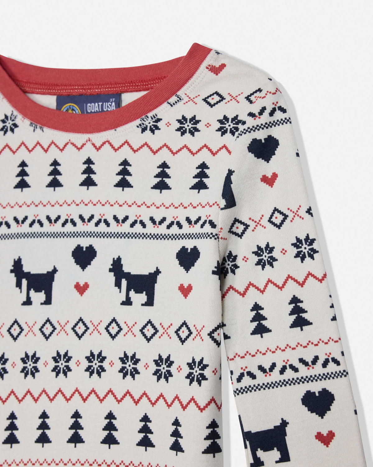 A close-up of a long-sleeve knit sweater with a cream background and a festive holiday pattern featuring black goats, red hearts, and red and black zigzag stripes. The inner collar shows a blue tag reading GOAT USA.