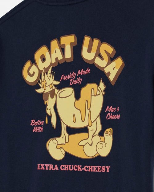 Close-up of a navy blue t-shirt featuring a retro graphic of a cartoon goat wearing sunglasses with the text 'GOAT USA' in large tan letters, 'Freshly Made Daily' in pink, and 'EXTRA CHUCK-CHEESY' at the bottom.