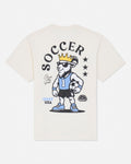 An off-white short-sleeve t-shirt with a large graphic on the back featuring a cartoon goat wearing a soccer jersey, shorts, and a gold crown under the word 'SOCCER'.