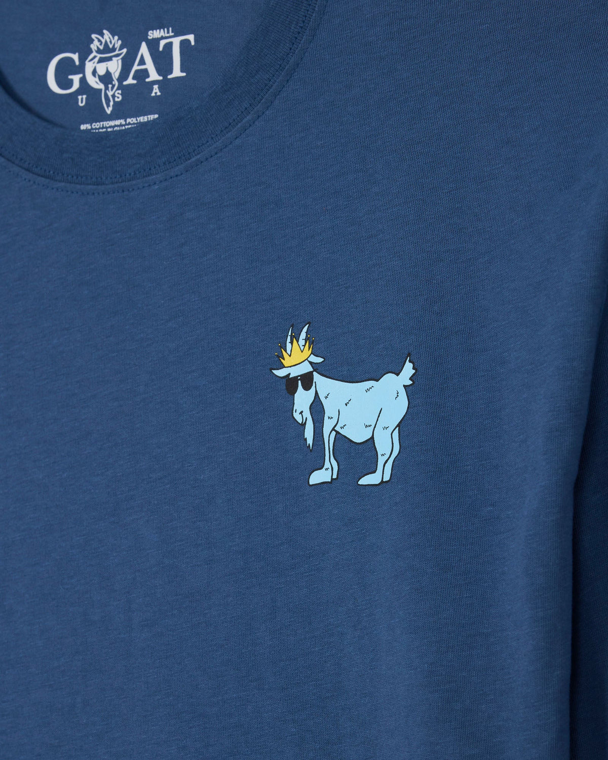 Close-up of a blue heathered t-shirt featuring a small graphic of a light blue goat wearing a gold crown and black sunglasses on the chest. The inner neck label displays the white 'GOAT' brand logo.