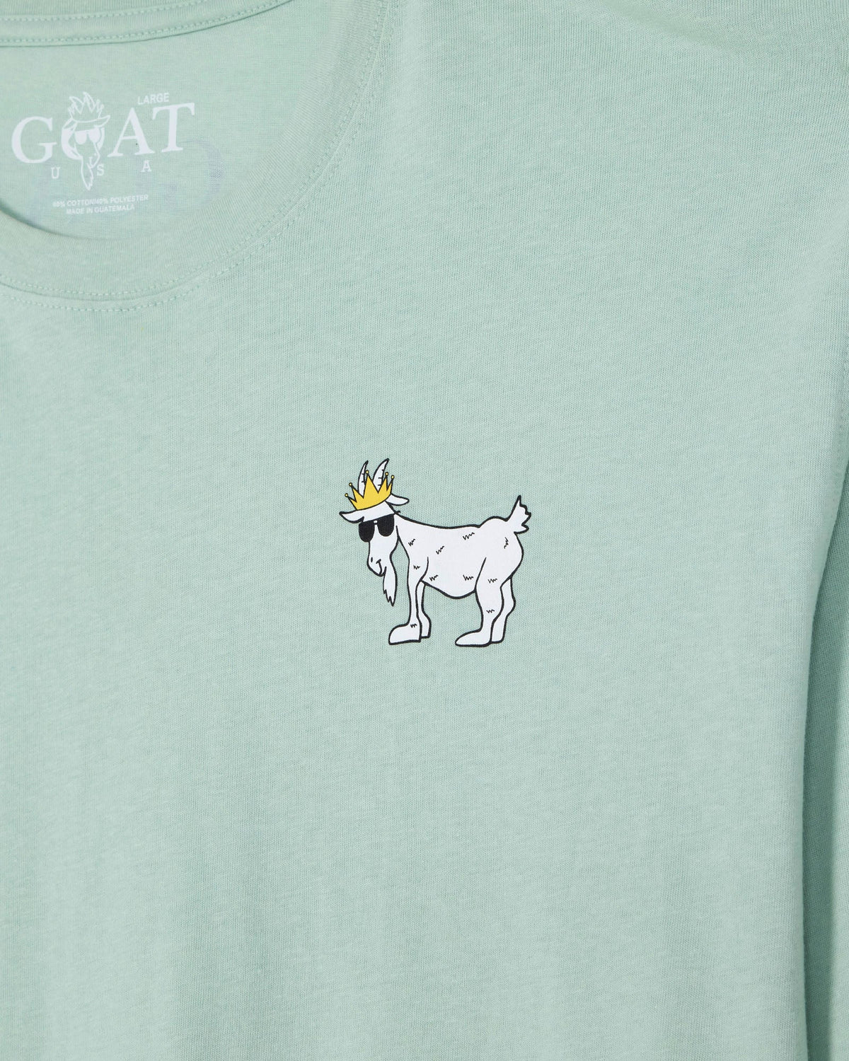 A close-up of a light mint green t-shirt featuring a small white cartoon goat wearing a gold crown and black sunglasses on the chest. The inner neck label shows the white printed 'GOAT' brand logo.