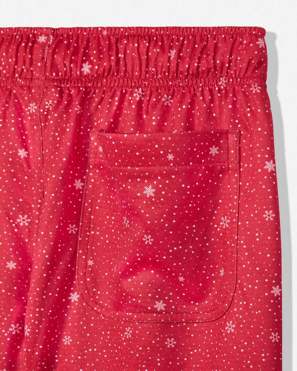 A close-up of red elastic-waist sleep pants featuring a white snowflake and star print, with a front patch pocket.