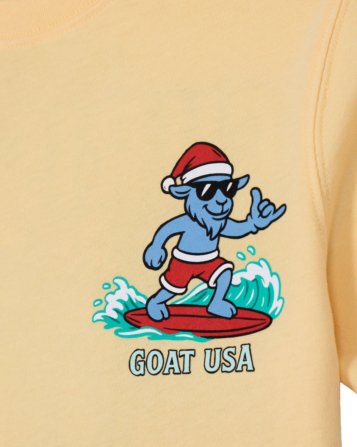 Close-up of a pale yellow t-shirt featuring a cartoon blue goat surfing on a red surfboard while wearing a red Santa hat and sunglasses, with the text 'GOAT USA' printed below.