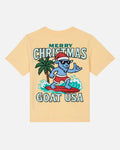 A pale yellow short-sleeve t-shirt with a colorful graphic on the front. The graphic features a blue cartoon goat wearing a red Santa hat and red swim trunks, surfing on a red surfboard next to a palm tree, with the text 'MERRY CHRISTMAS GOAT USA' in stylized lettering.
