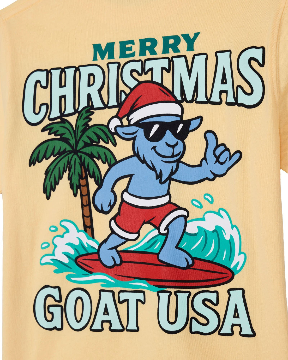 A close-up of a light beige t-shirt featuring a colorful graphic print. The design shows a blue cartoon goat wearing a red Santa hat and sunglasses, surfing on a red surfboard with a green palm tree and white waves behind it.