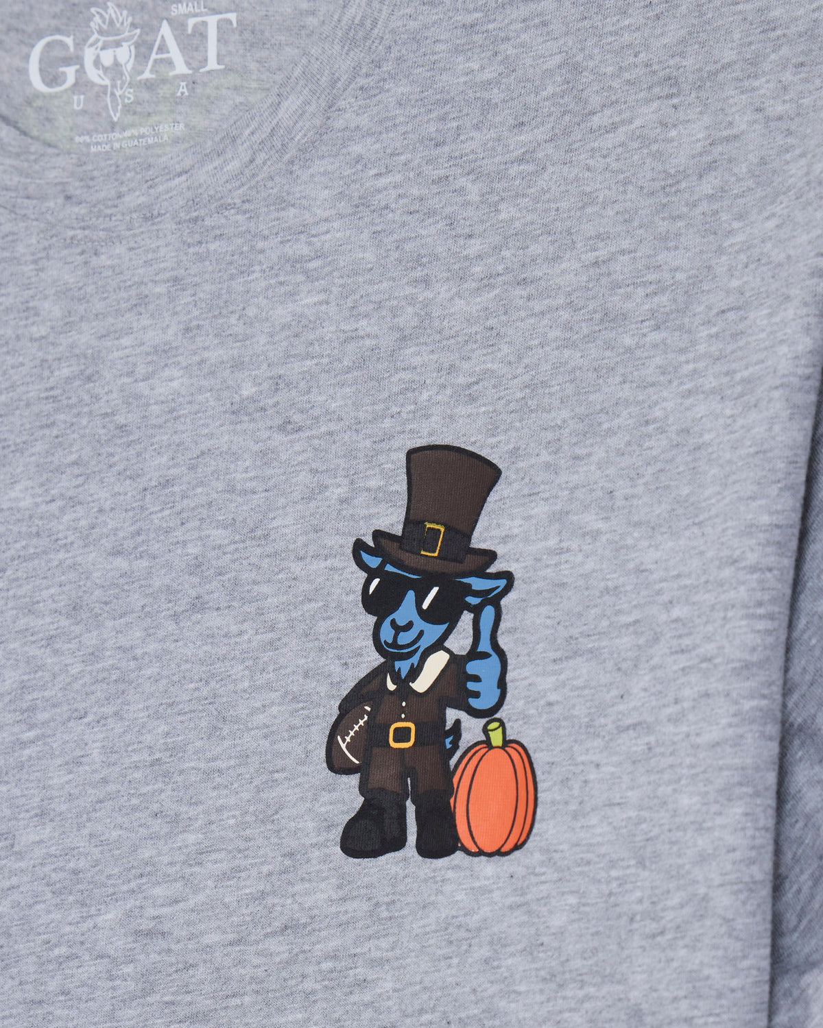 Close-up of a heather gray t-shirt featuring a cartoon blue goat character dressed as a Thanksgiving pilgrim with a black top hat, holding an orange pumpkin.
