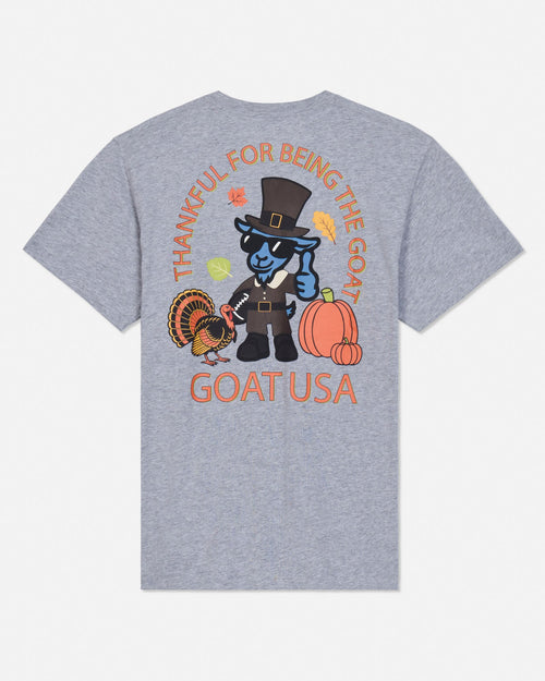 A heather gray short-sleeve t-shirt shown from the back, featuring a colorful graphic of a blue goat dressed as a pilgrim with pumpkins, a turkey, and autumn leaves, with the text 'THANKFUL FOR BEING THE GOAT' and 'GOATUSA'.