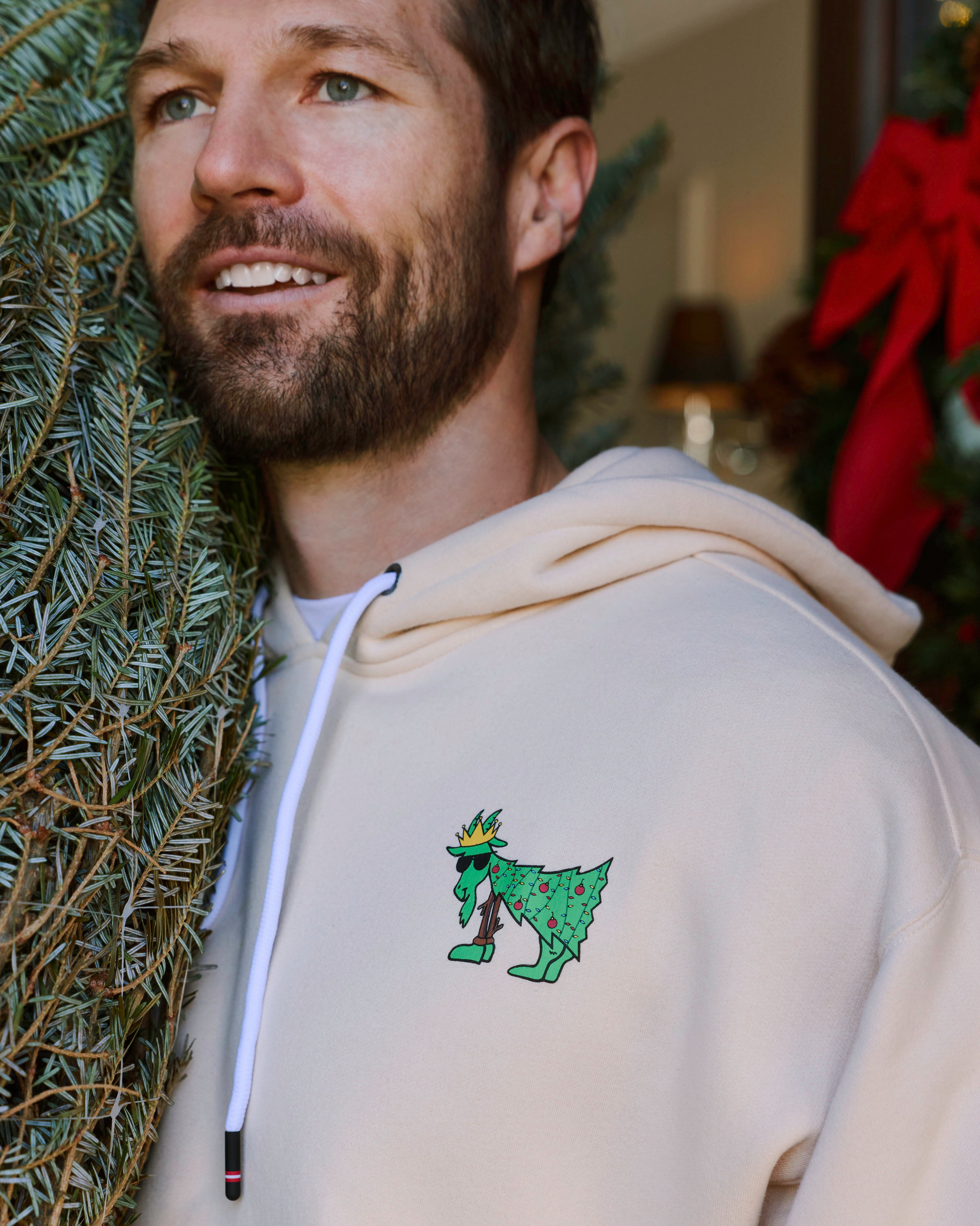 Man wearing a beige hoodie with a green graphic design, standing next to a Christmas tree.