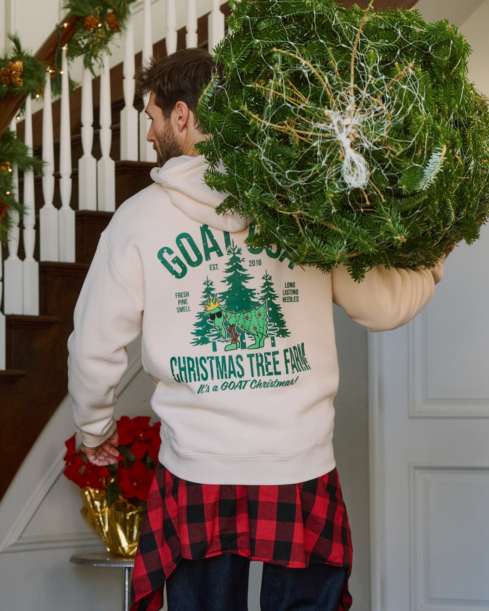 Person carrying a Christmas tree wearing a hoodie with a Christmas tree farm design.