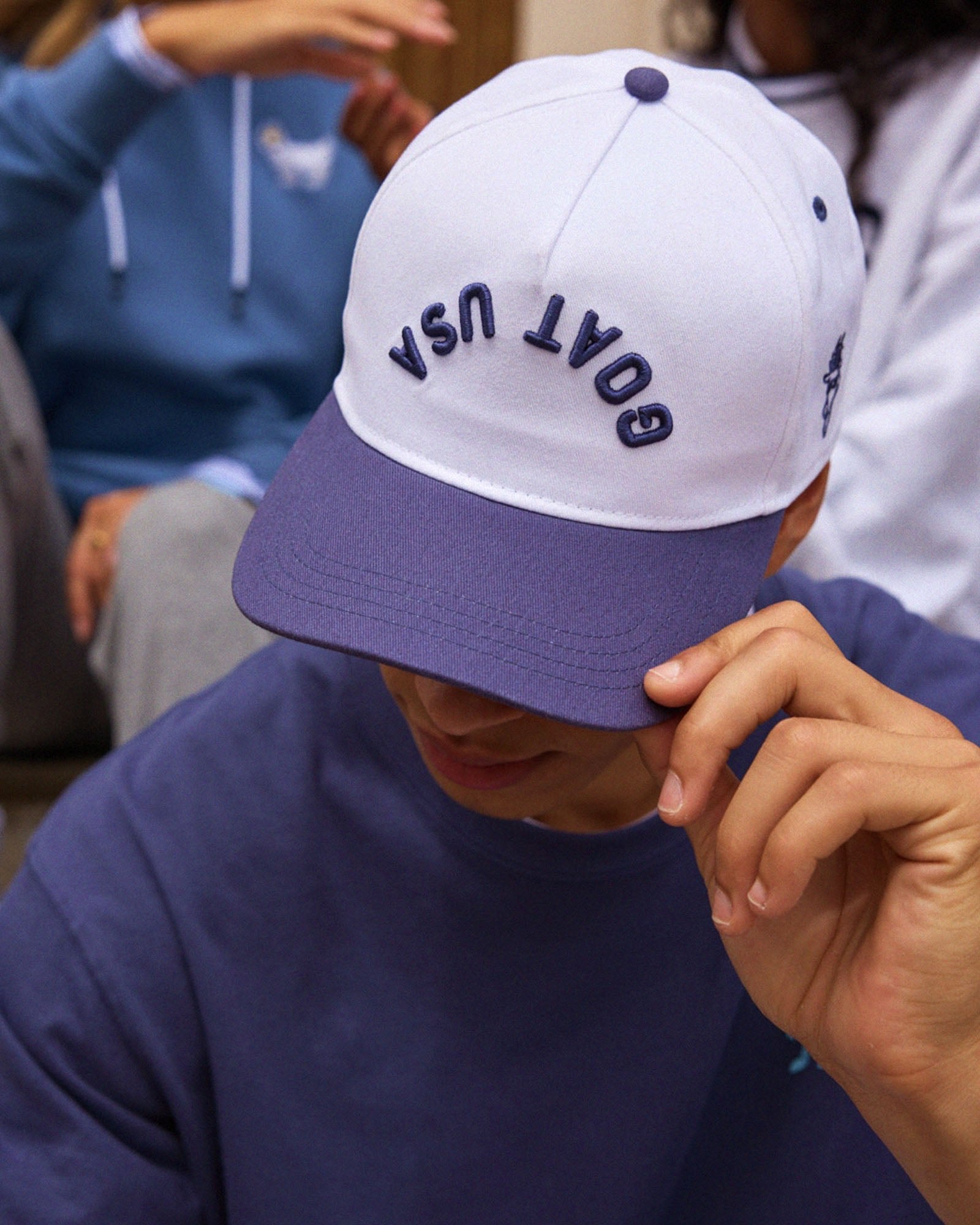Person wearing a white and blue cap with 'GOAT USA' upside down text, sitting among others.