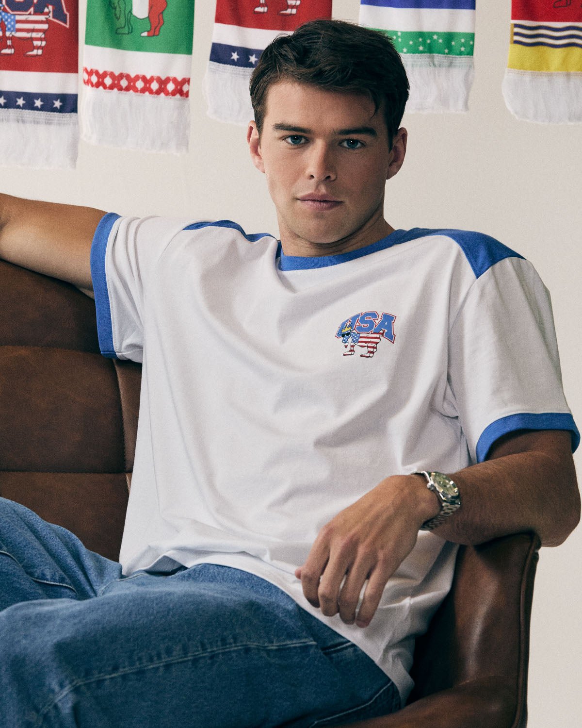 Man wearing a white t-shirt with blue accents and 'USA' logo, sitting in front of flags.