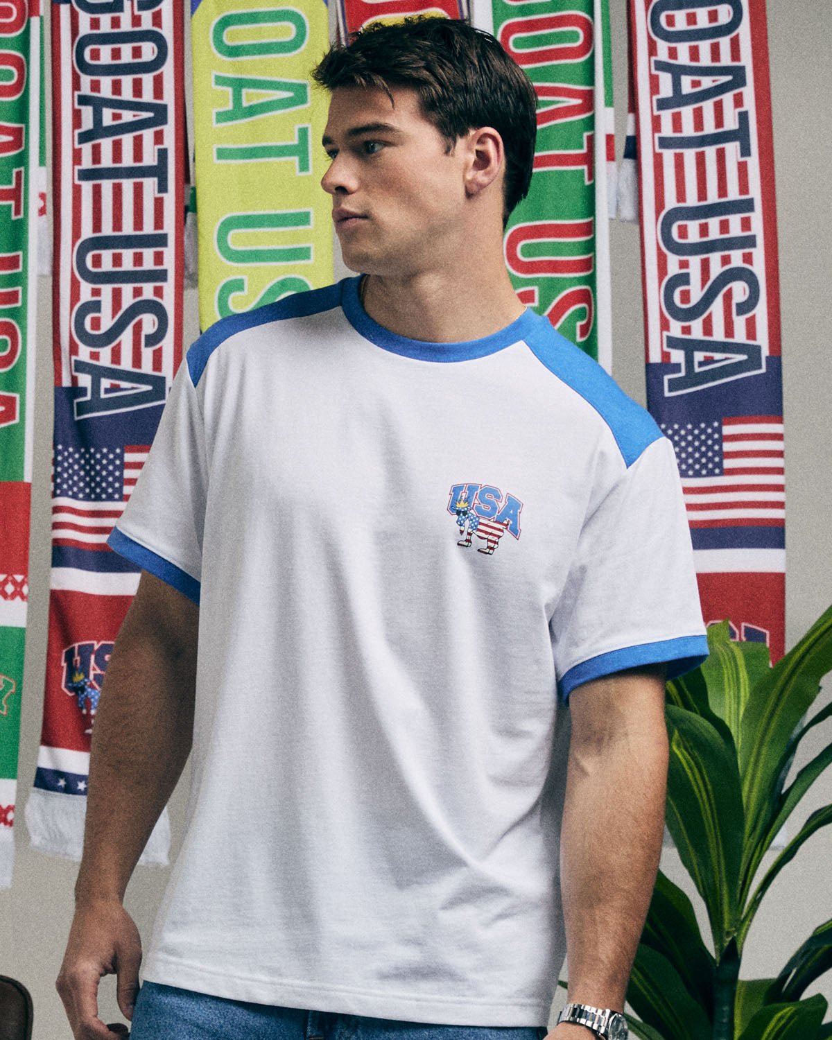 Man wearing a white t-shirt with blue sleeves and 'USA' logo in front of colorful flags.