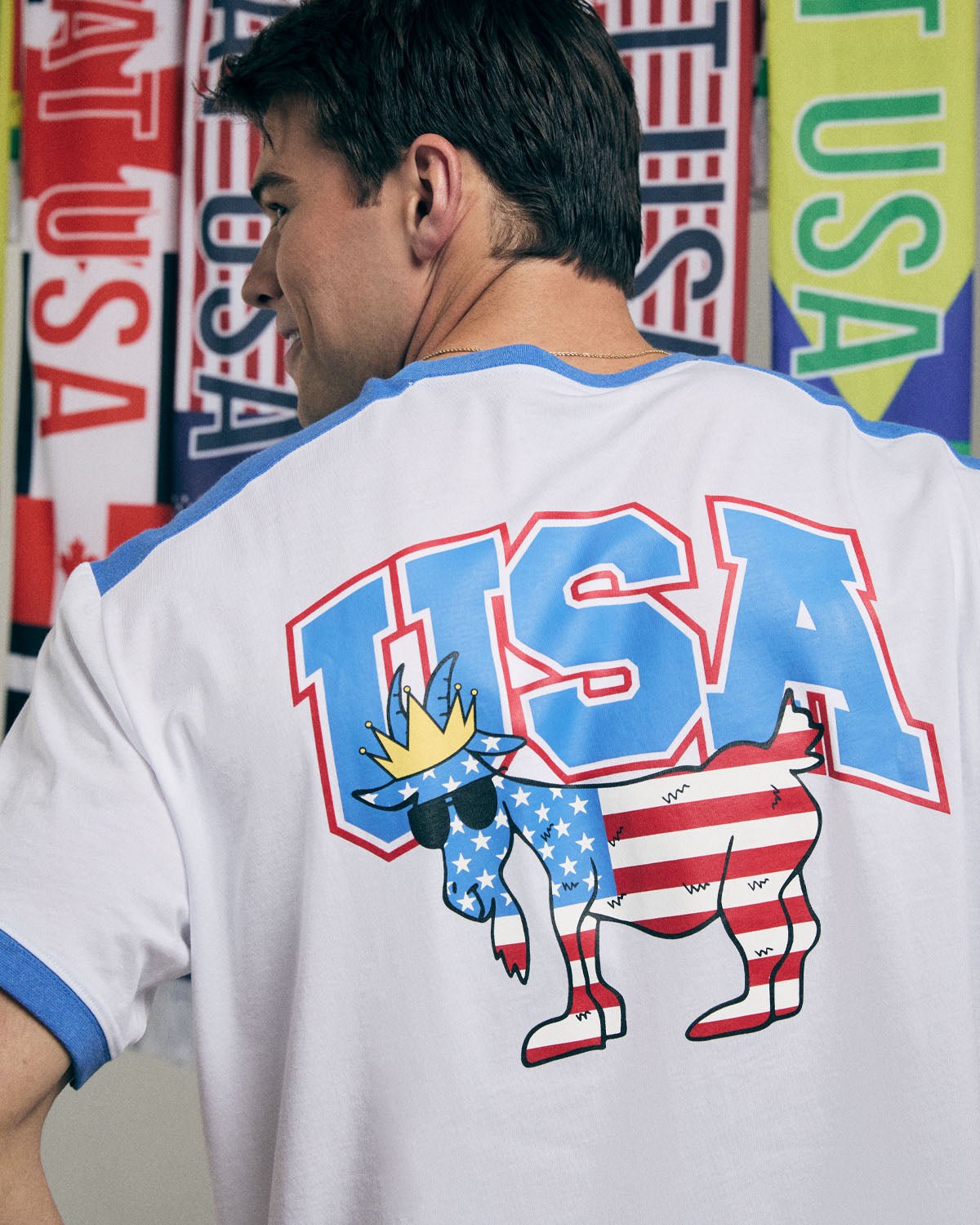 Person wearing a white t-shirt with 'USA' and goat graphic in front of USA flags.