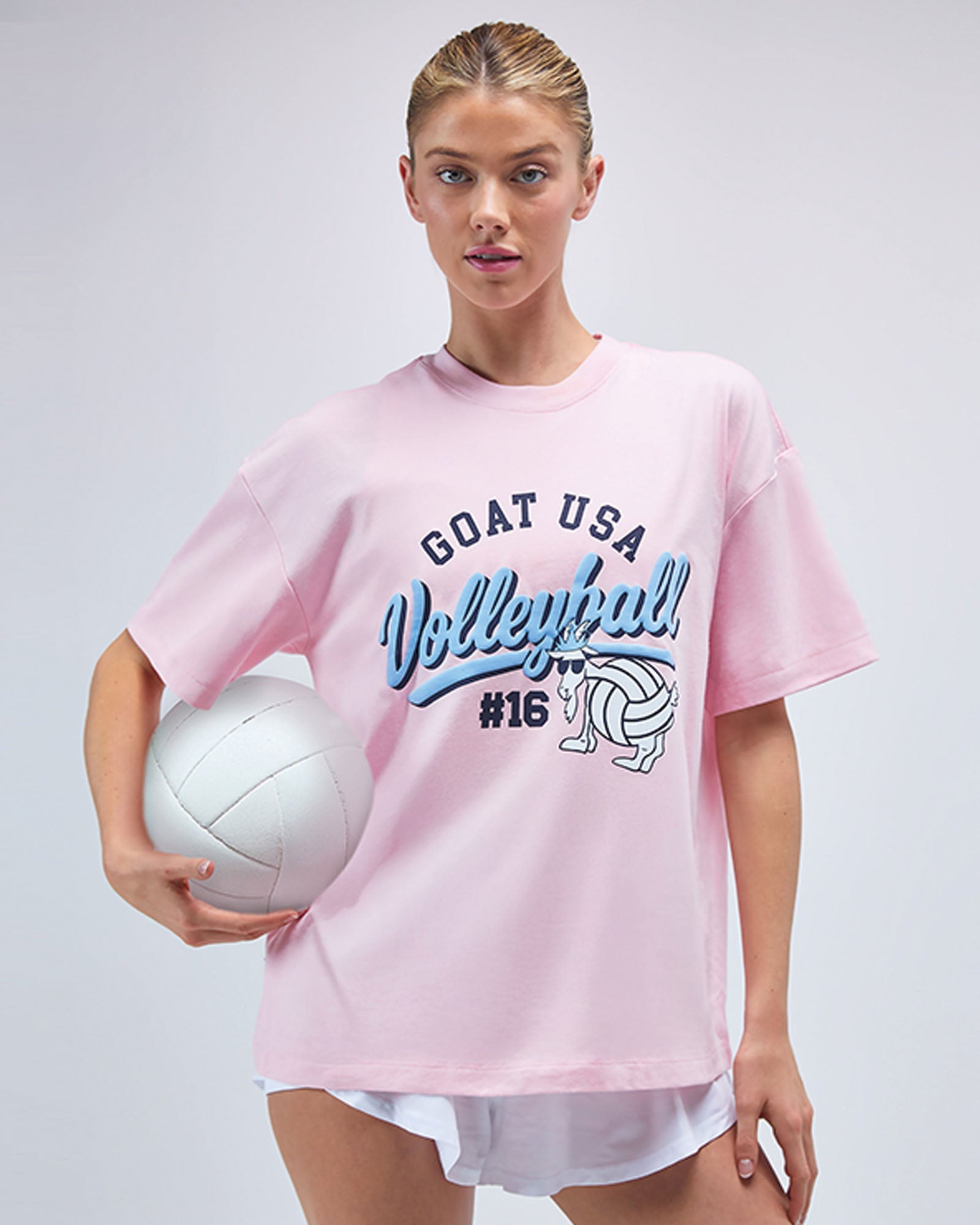 Woman wearing a pink 'GOAT USA Volleyball #16' t-shirt holding a volleyball on a white background