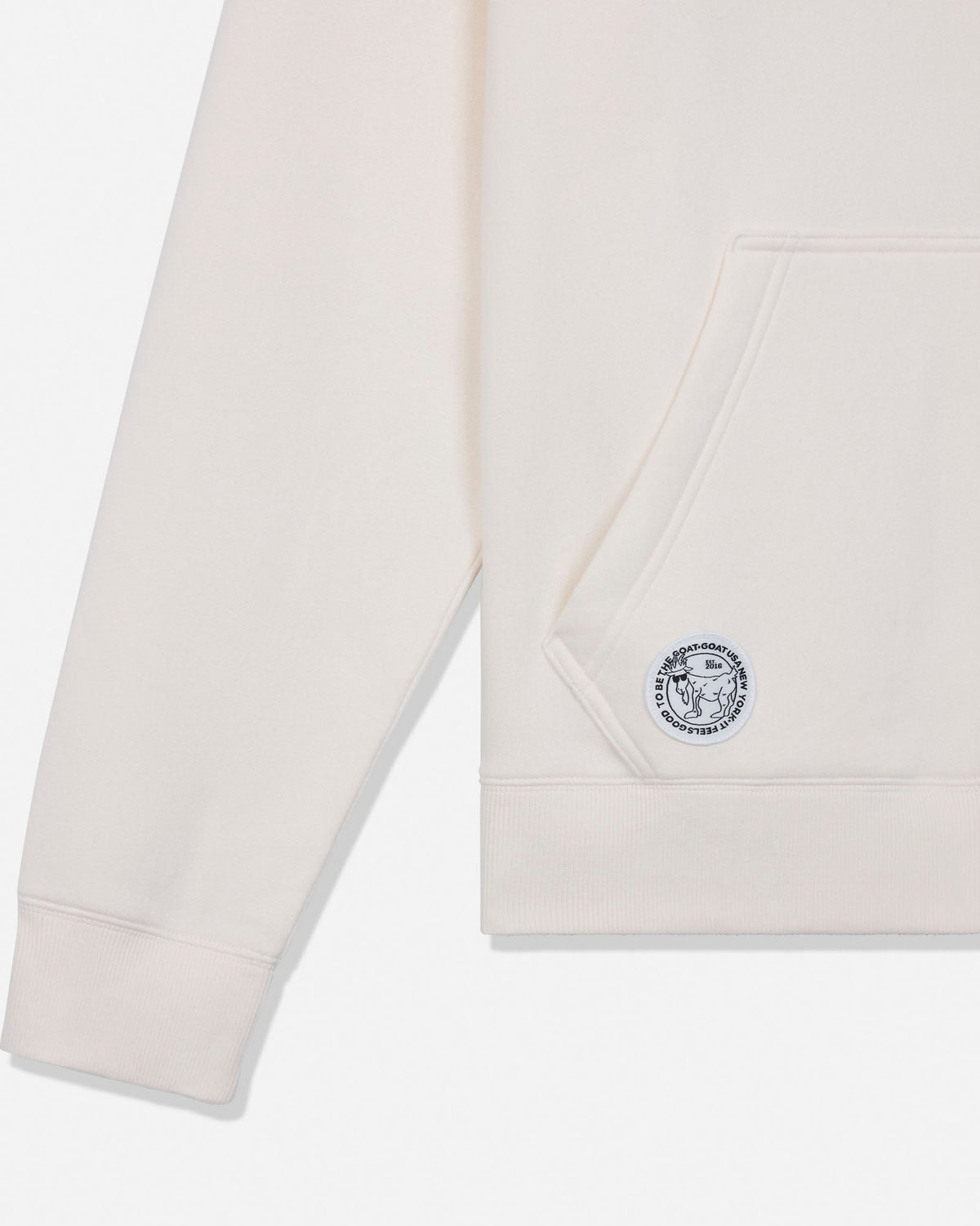 Close-up of a light cream-colored hoodie showing the sleeve, pocket, and a circular embroidered logo patch on the pocket.