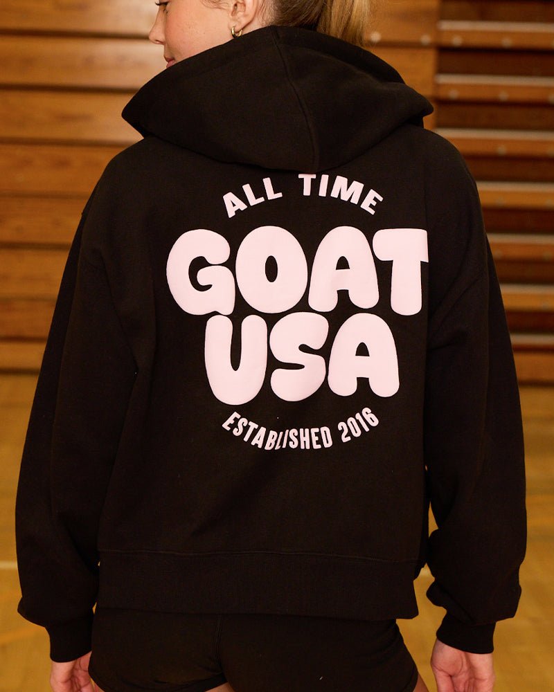 Back of black full-zip hoodie with pink 'GOAT USA' on a female model#color_black