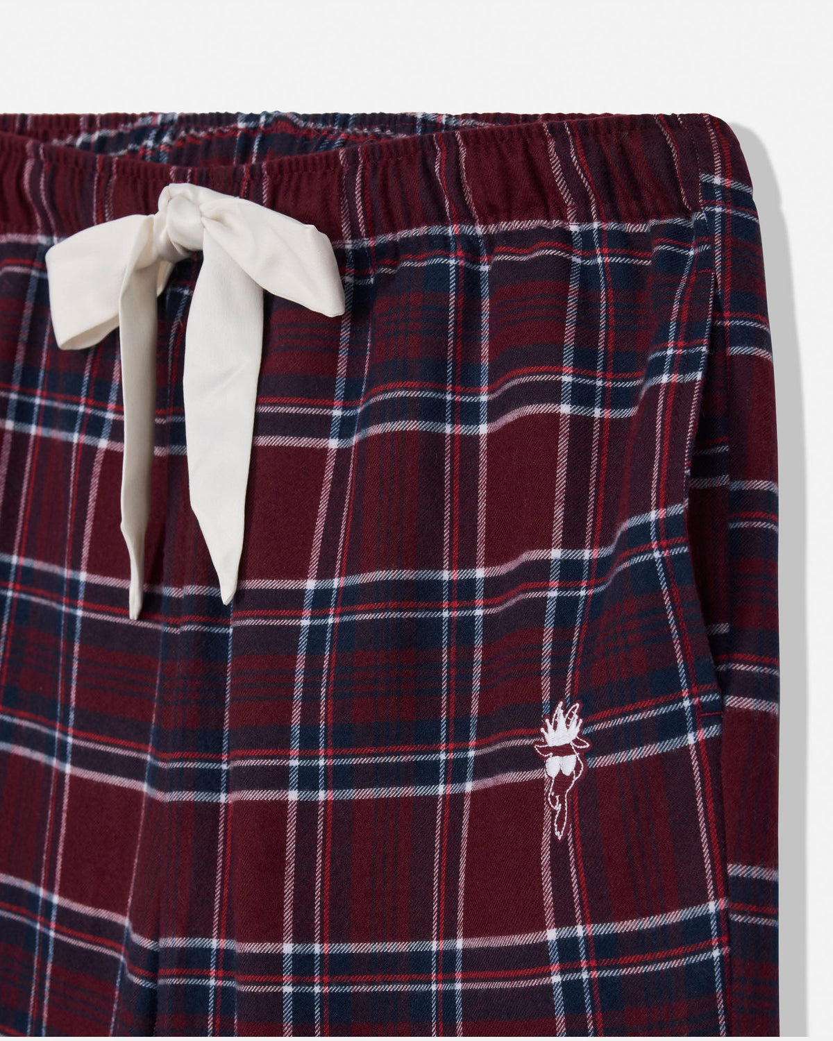 Close-up of a plaid lounge or pajama bottom in deep burgundy with navy and white stripes, featuring an off-white drawstring bow and a small embroidered goat detail on the lower right leg.