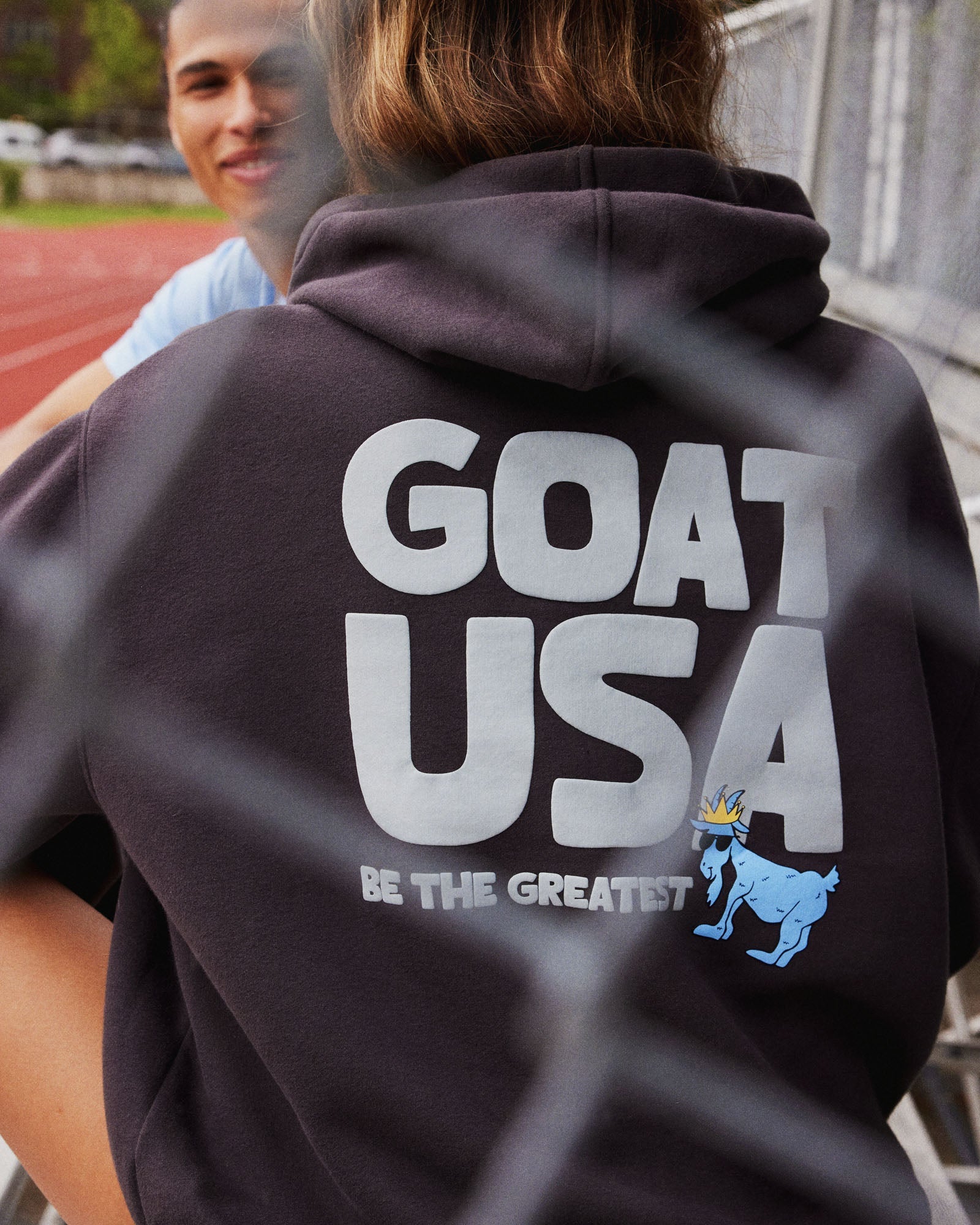 Person wearing a hoodie with 'GOAT USA' text and logo, standing behind a chain-link fence.#color_dark-gray