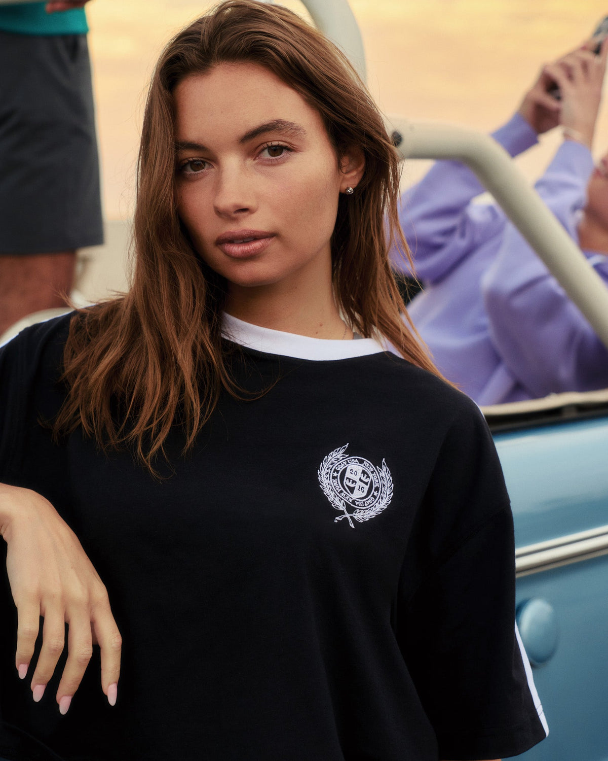 Woman wearing a black tee with a logo, standing in front of a vintage car.