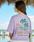Girl wearing a purple top with palm tree design with water and a dock in the background