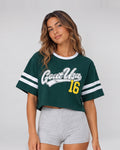 A woman modeling a dark green cropped t-shirt with 'Goat Usa 16' printed on the front, featuring white and gray athletic stripes on the sleeves, paired with light gray shorts against a light gray studio background.