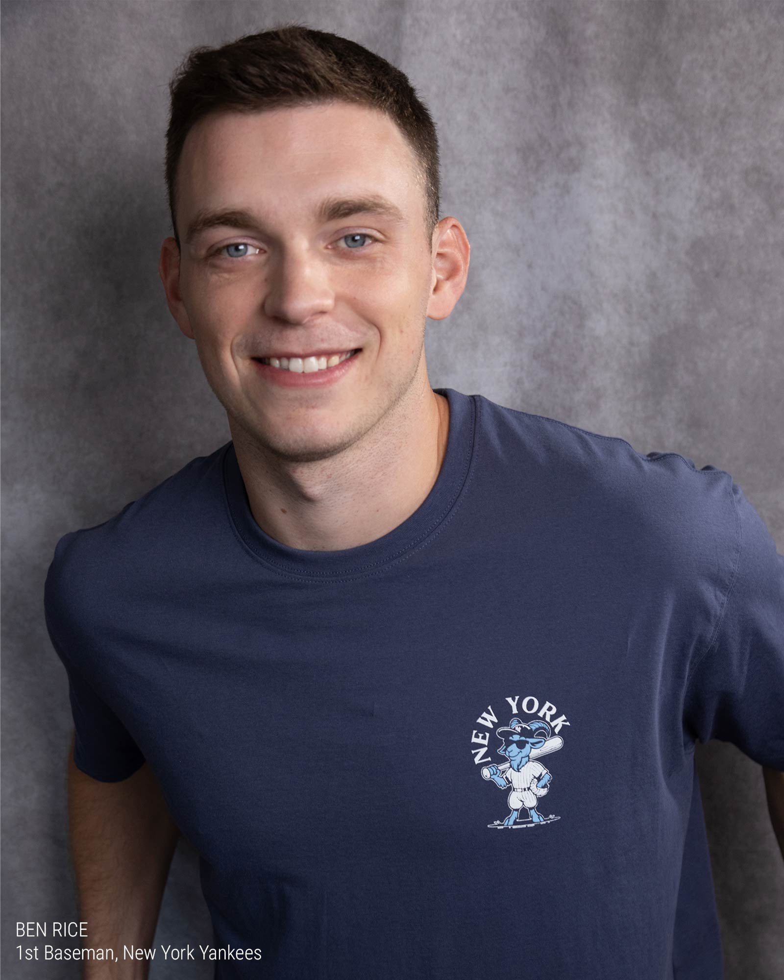 A smiling man wearing a navy blue crewneck t-shirt with a white and blue New York Yankees graphic on the left chest, set against a textured gray studio background.