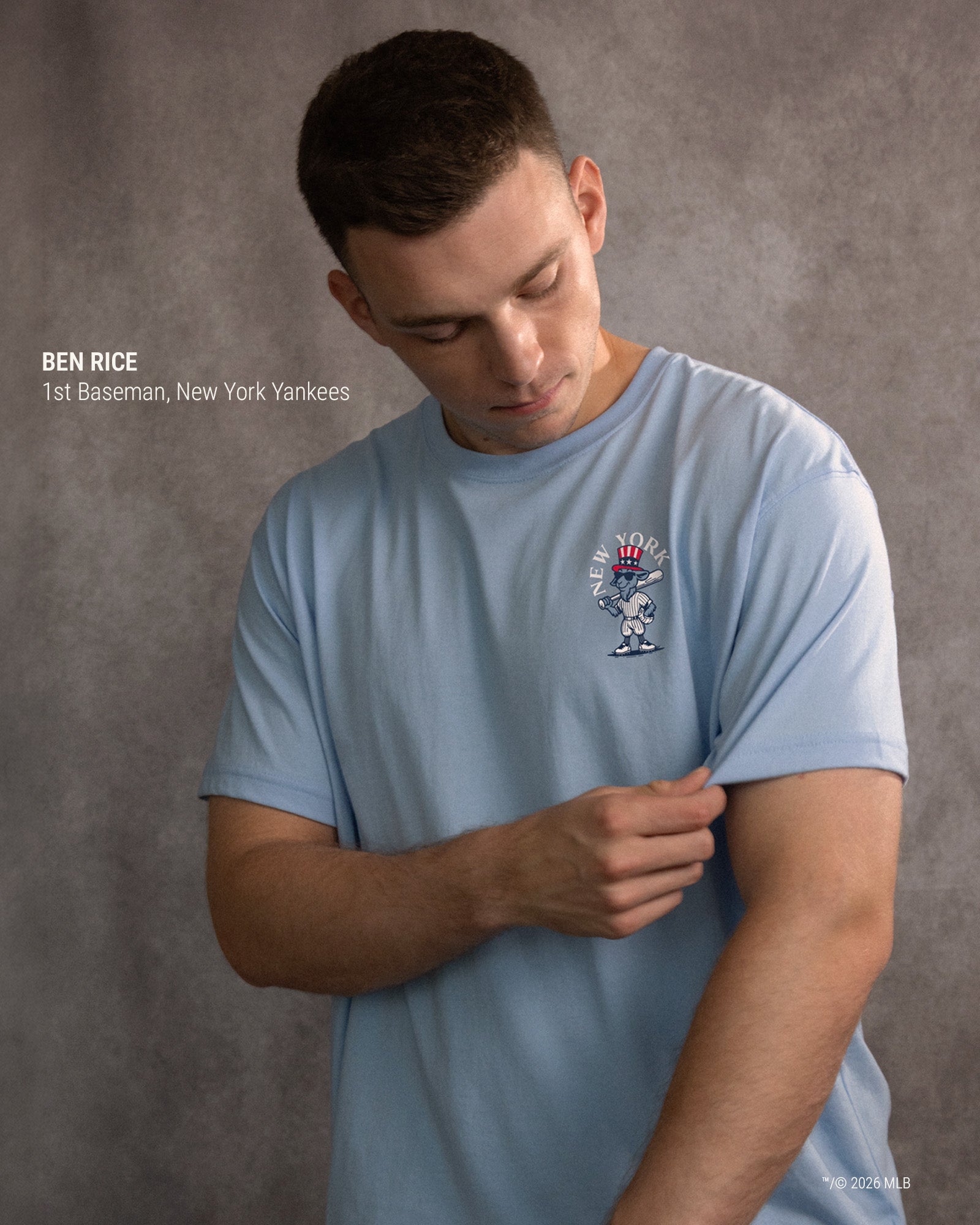 A man wearing a light blue crewneck t-shirt with a New York Yankees baseball graphic on the left chest, shown against a textured gray studio background.#color_carolina-blue