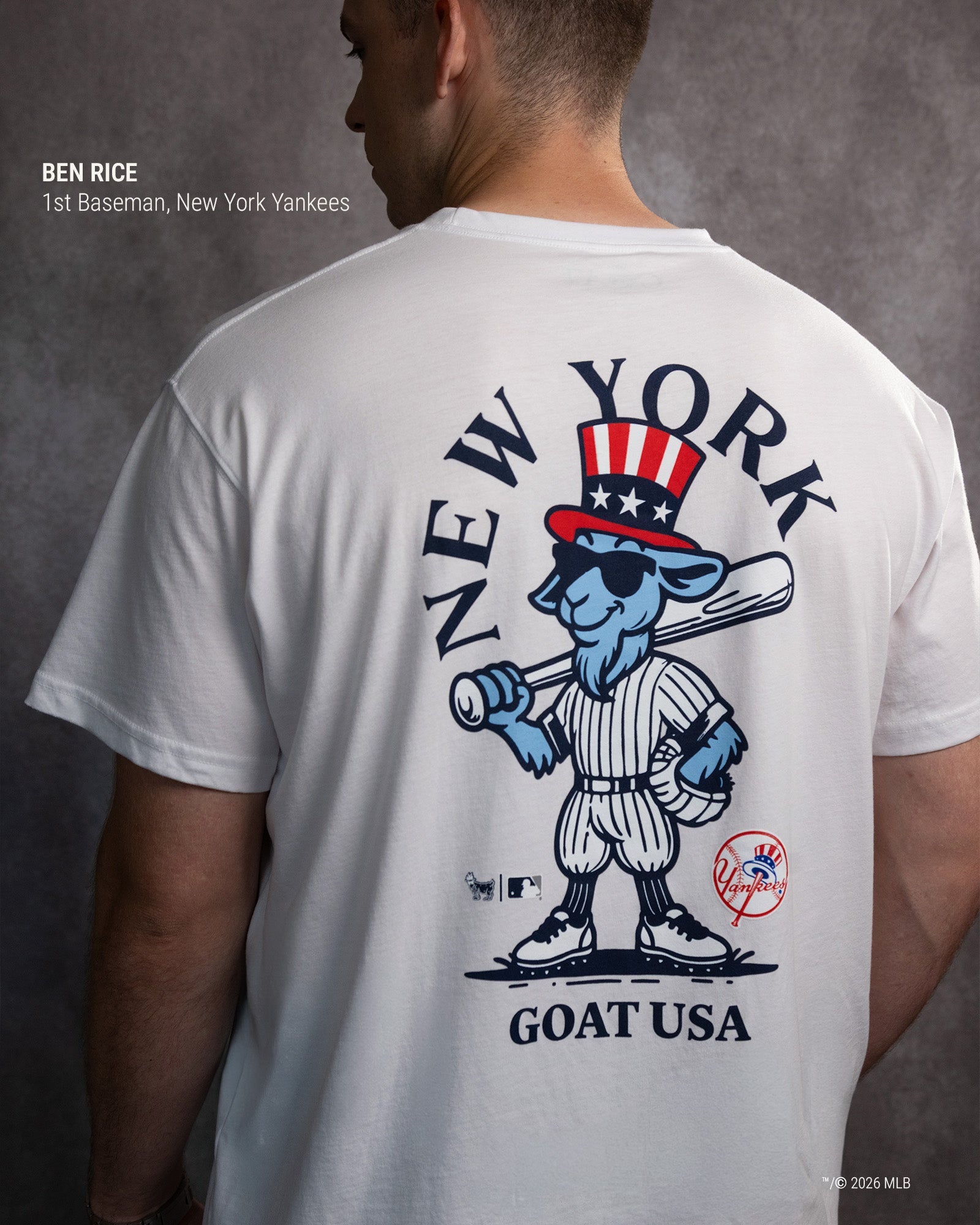 A man shown from behind wearing a white t-shirt with a large back graphic. The graphic features a blue cartoon goat in a New York Yankees patriotic Uncle Sam hat and pinstripe uniform, with 'NEW YORK' arched above and 'GOAT USA' below.#color_white